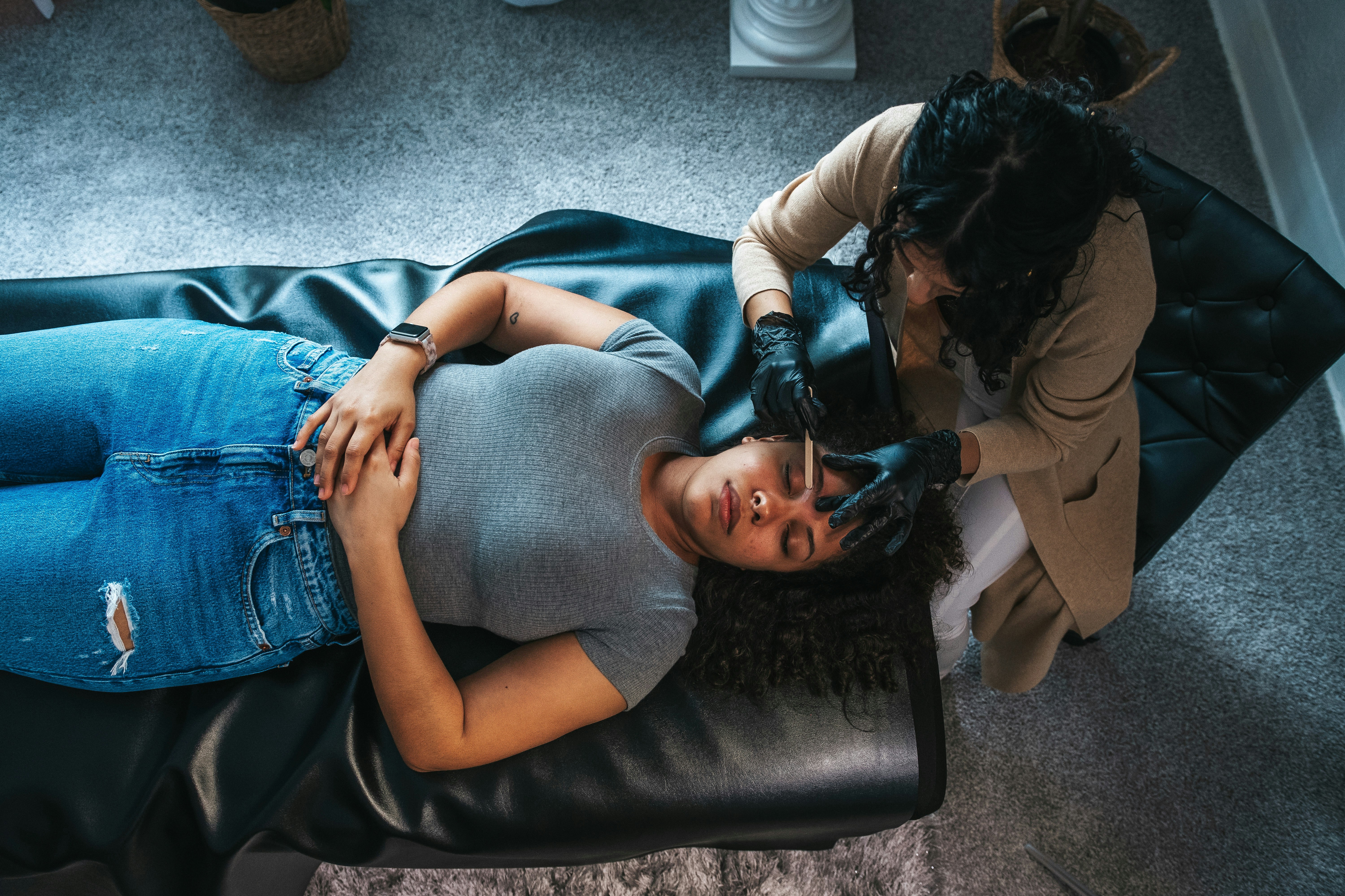 An aesthetician in black gloves shaping a client’s brows, seen from above